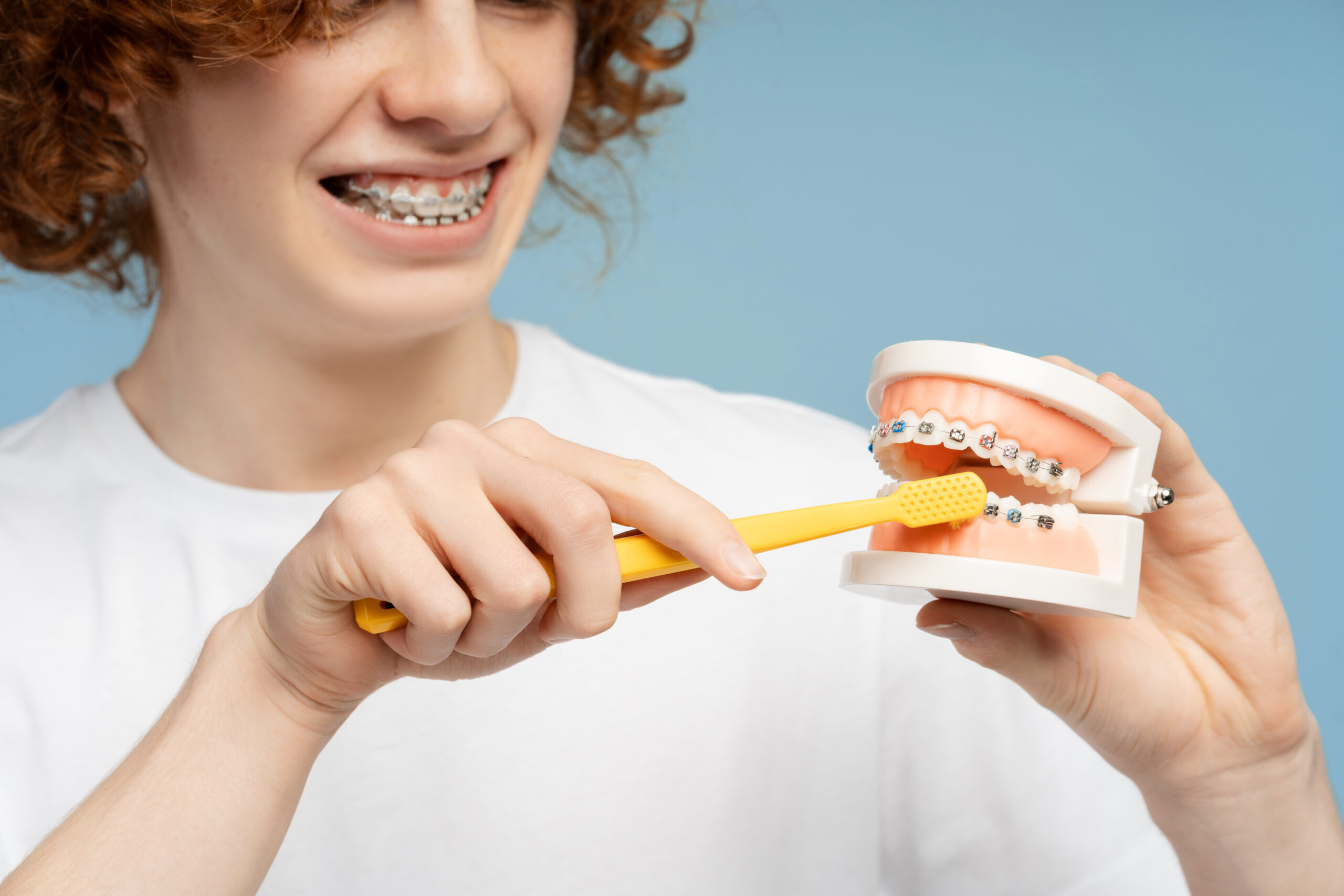 Smiling, curly red headed teen male with braces, close up photo, brushing a dental model, isolated on a blue background. Focus on dental hygiene concept